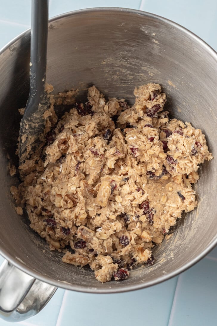 A bowl of cookie dough with oats and dried cranberries, mixed with a black spatula, resting on a light blue countertop, creating a cozy, homemade feel.