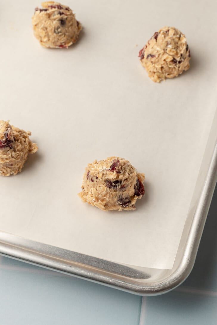 Cookie dough scoops with oats and cranberries on a parchment-lined baking sheet, ready for baking. The scene suggests anticipation and warmth.