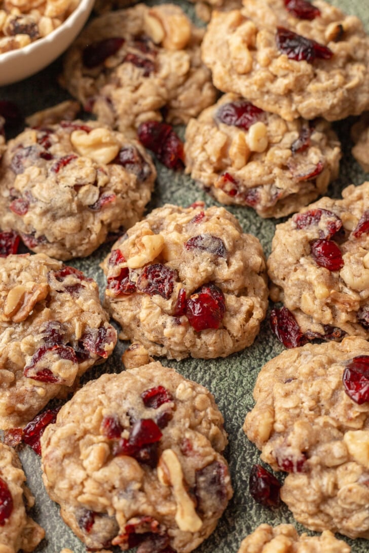 Close-up of oatmeal cookies with cranberries and nuts on a textured surface. The cookies appear golden and chunky, conveying a homemade feel.