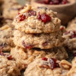 A stack of oatmeal cookies with cranberries and walnuts on a textured surface. The cookies are golden brown and rustic, evoking a warm, homemade feel.