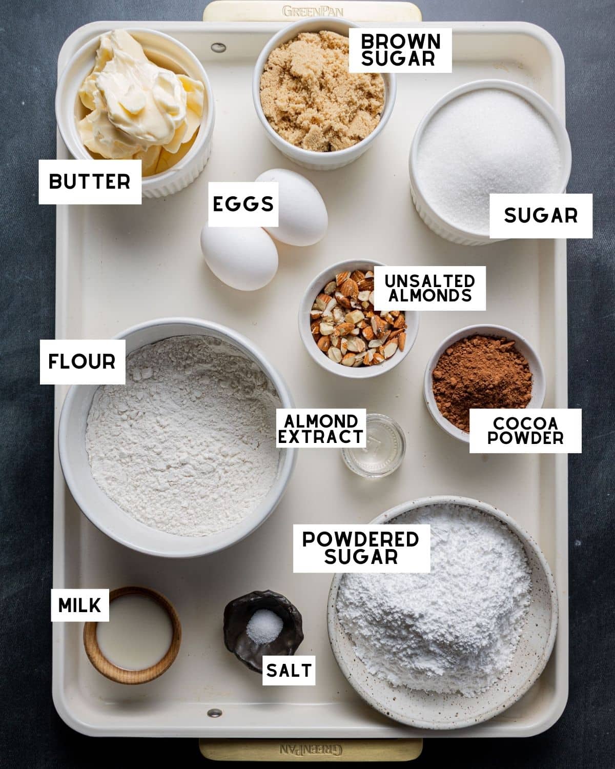 A baking tray displays labeled ingredients: butter, eggs, brown sugar, sugar, flour, unsalted almonds, cocoa powder, almond extract, powdered sugar, milk, and salt.