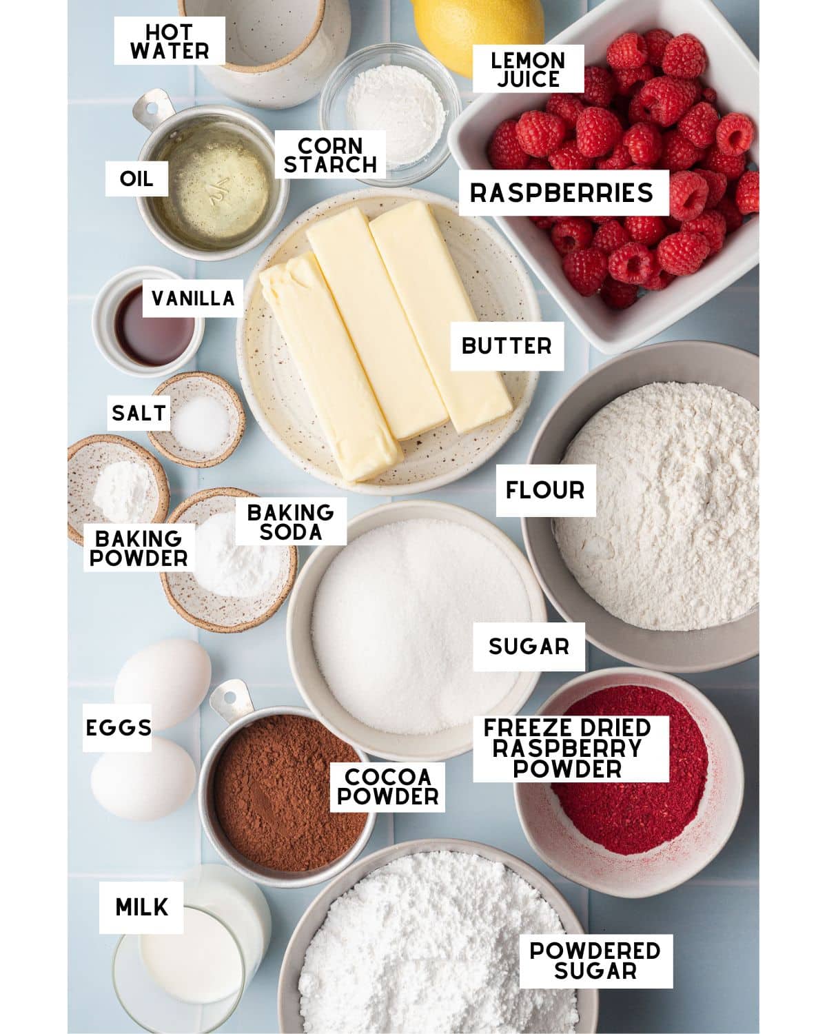 A neatly arranged assortment of baking ingredients includes raspberries, flour, butter, eggs, sugar, cocoa powder, and milk, labeled for clarity. The image conveys a sense of organization and preparation for baking.