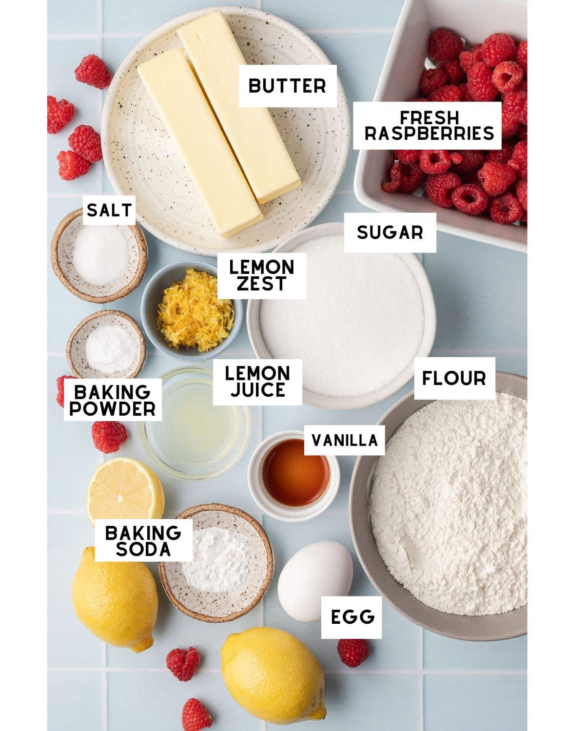 Overhead view of dessert ingredients on a blue surface. Items include butter, fresh raspberries, sugar, flour, egg, lemon, and vanilla.