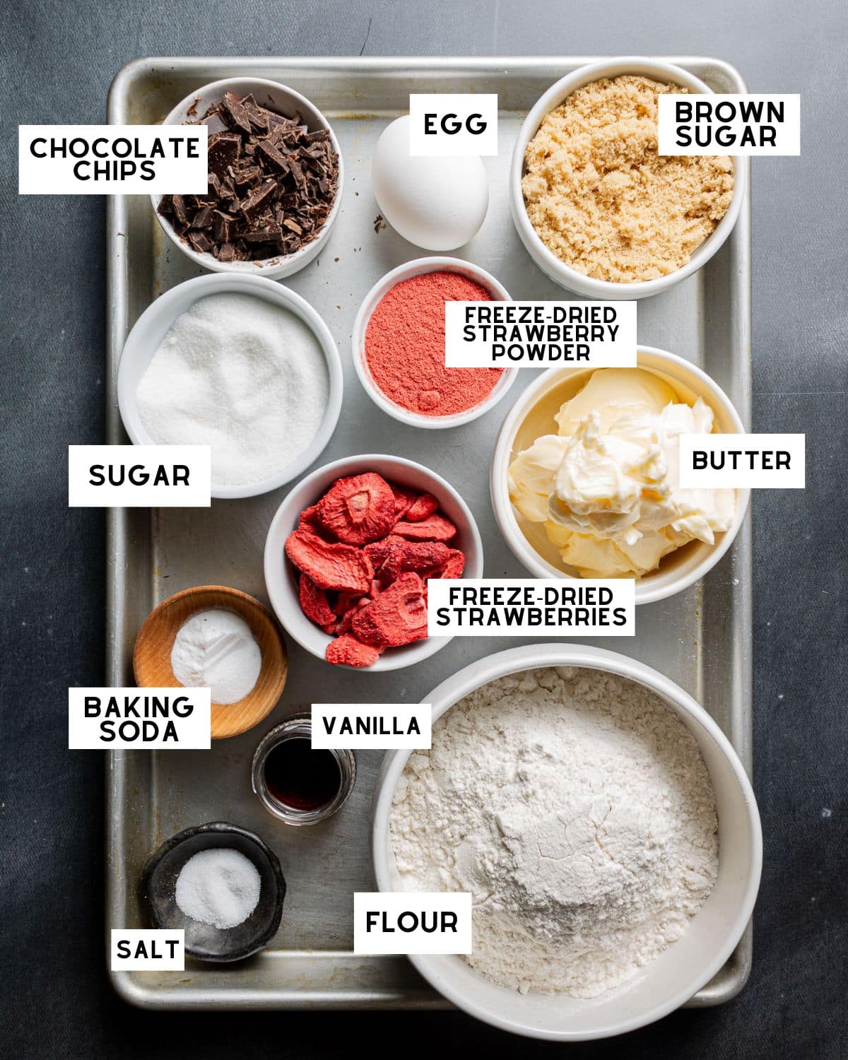 Baking sheet with labeled ingredients: egg, brown sugar, butter, chocolate chips, sugar, freeze-dried strawberries, strawberry powder, flour, vanilla, baking soda, salt.