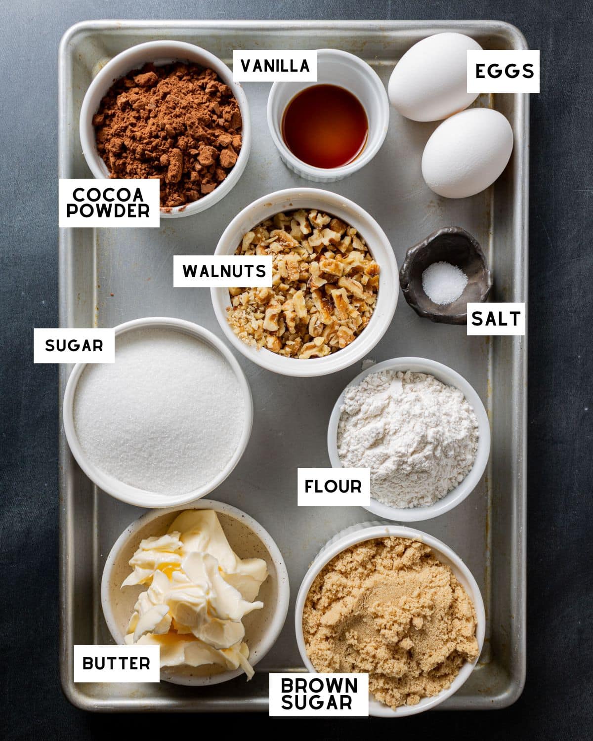 A baking tray displays labeled ingredients: cocoa powder, vanilla, eggs, walnuts, sugar, salt, flour, brown sugar, and butter, evoking a cozy baking atmosphere.