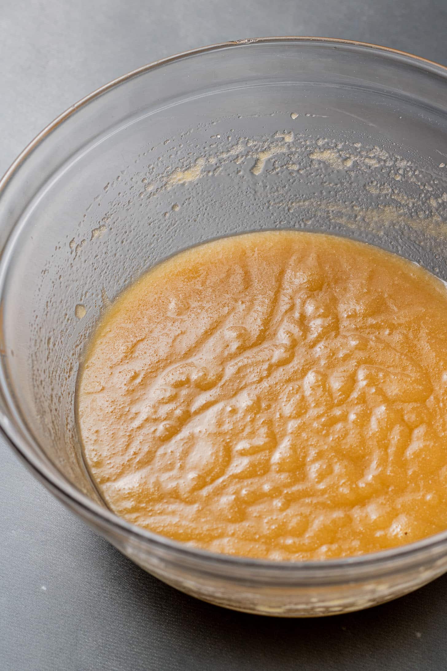 A glass bowl containing smooth, tan batter rests on a gray countertop. The batter's glossy surface suggests it is freshly mixed and ready for baking.