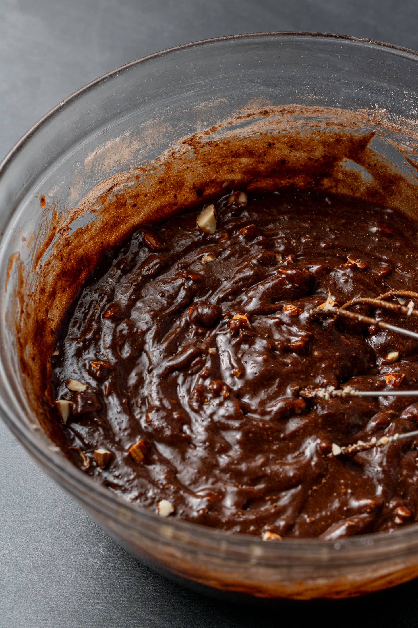 Close-up of a glass bowl filled with rich, dark chocolate brownie batter, featuring visible chunks of nuts and a smooth, creamy texture.