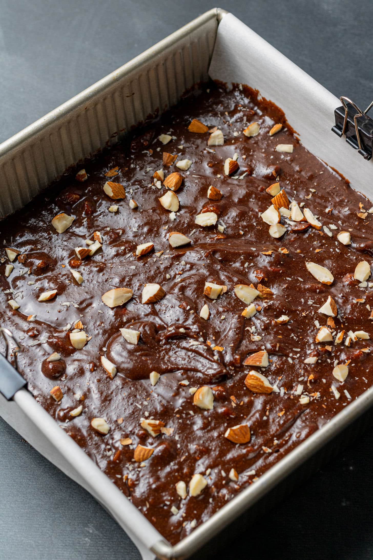 Chocolate brownie batter spread in a square baking pan, topped with chopped almonds. The smooth batter surface gives a homemade, inviting feel.