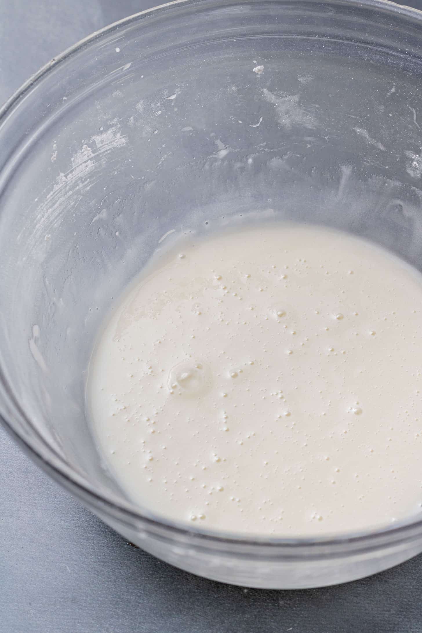 A clear glass bowl holds a pale, slightly bubbly glaze on a smooth gray surface, conveying a sense of homemade preparation and anticipation.