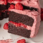 A chocolate cake with two layers of dark sponge and vibrant pink raspberry frosting. Fresh raspberries garnish the top. A slice is removed, exposing the rich texture.