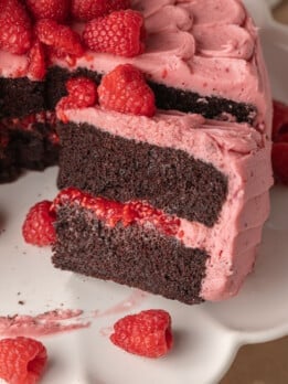 A chocolate cake with two layers of dark sponge and vibrant pink raspberry frosting. Fresh raspberries garnish the top. A slice is removed, exposing the rich texture.