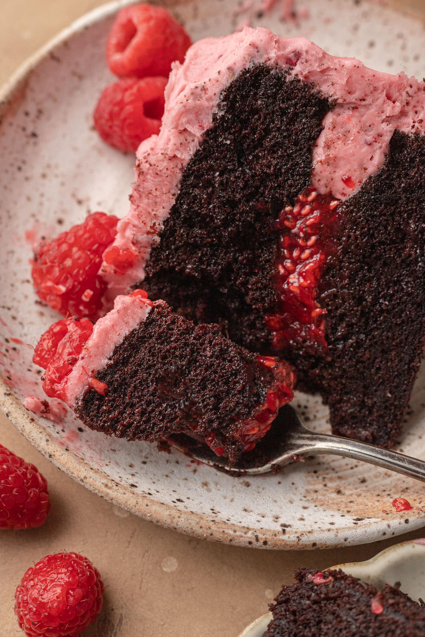 A slice of rich chocolate cake with pink raspberry frosting on a plate, topped with fresh raspberries. A fork holds a moist bite, creating a tempting scene.
