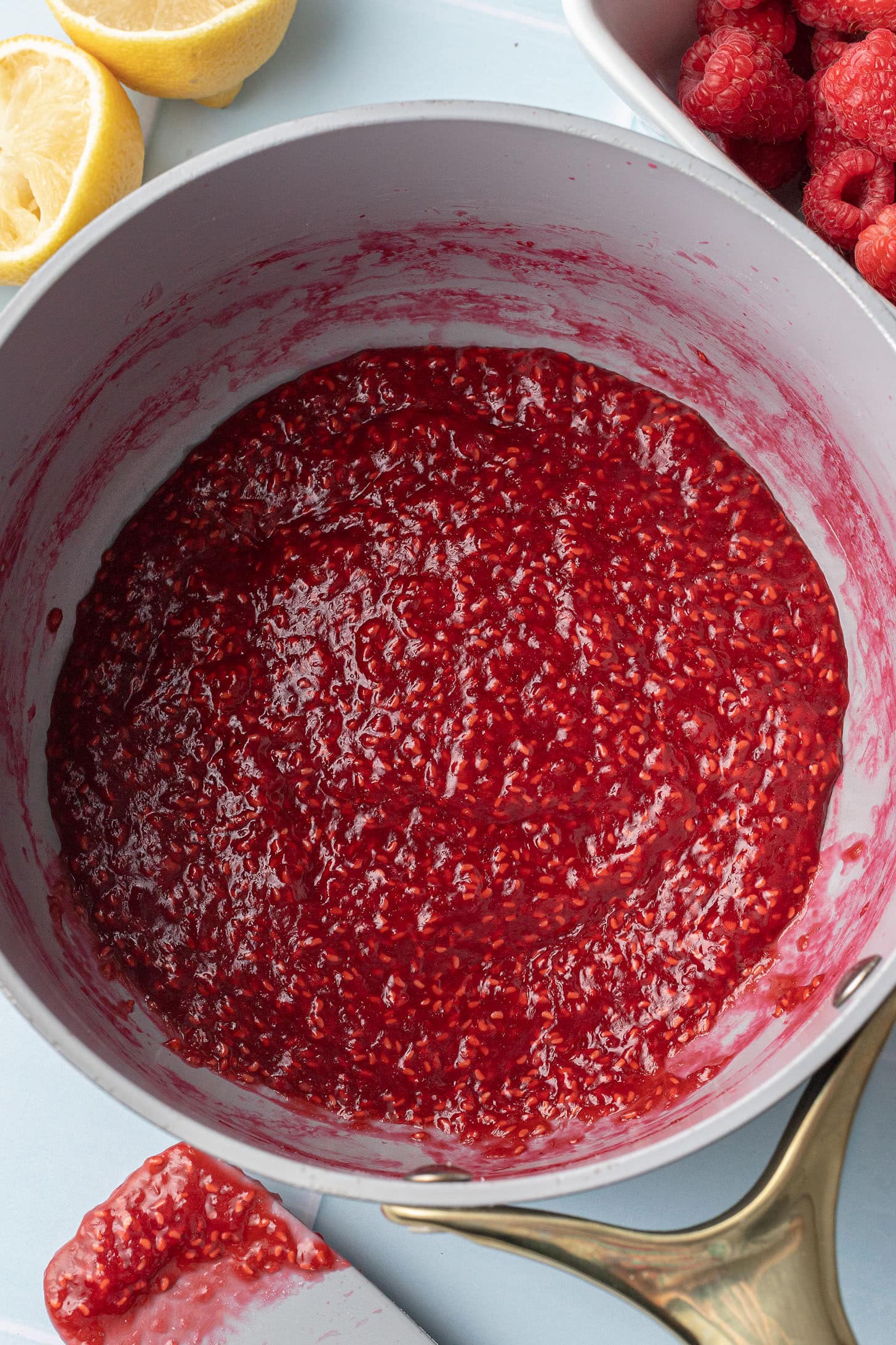 A pot of vibrant red raspberry sauce with seeds is shown, surrounded by fresh lemons and raspberries, conveying a fresh and zesty cooking scene.