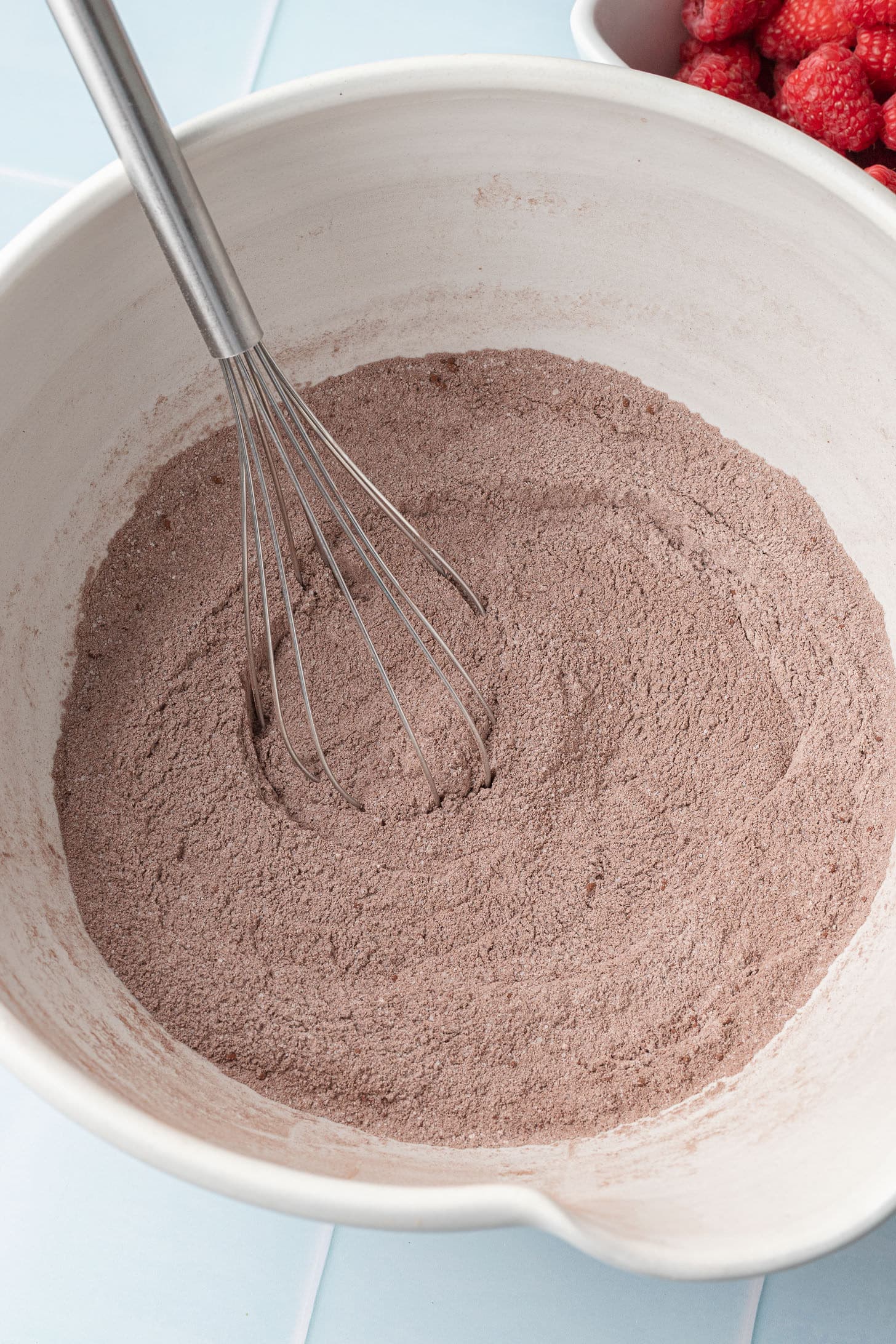 A white bowl with cocoa powder and flour being whisked. A whisk rests in the mixture. Fresh raspberries are visible in a bowl nearby, creating a baking scene.
