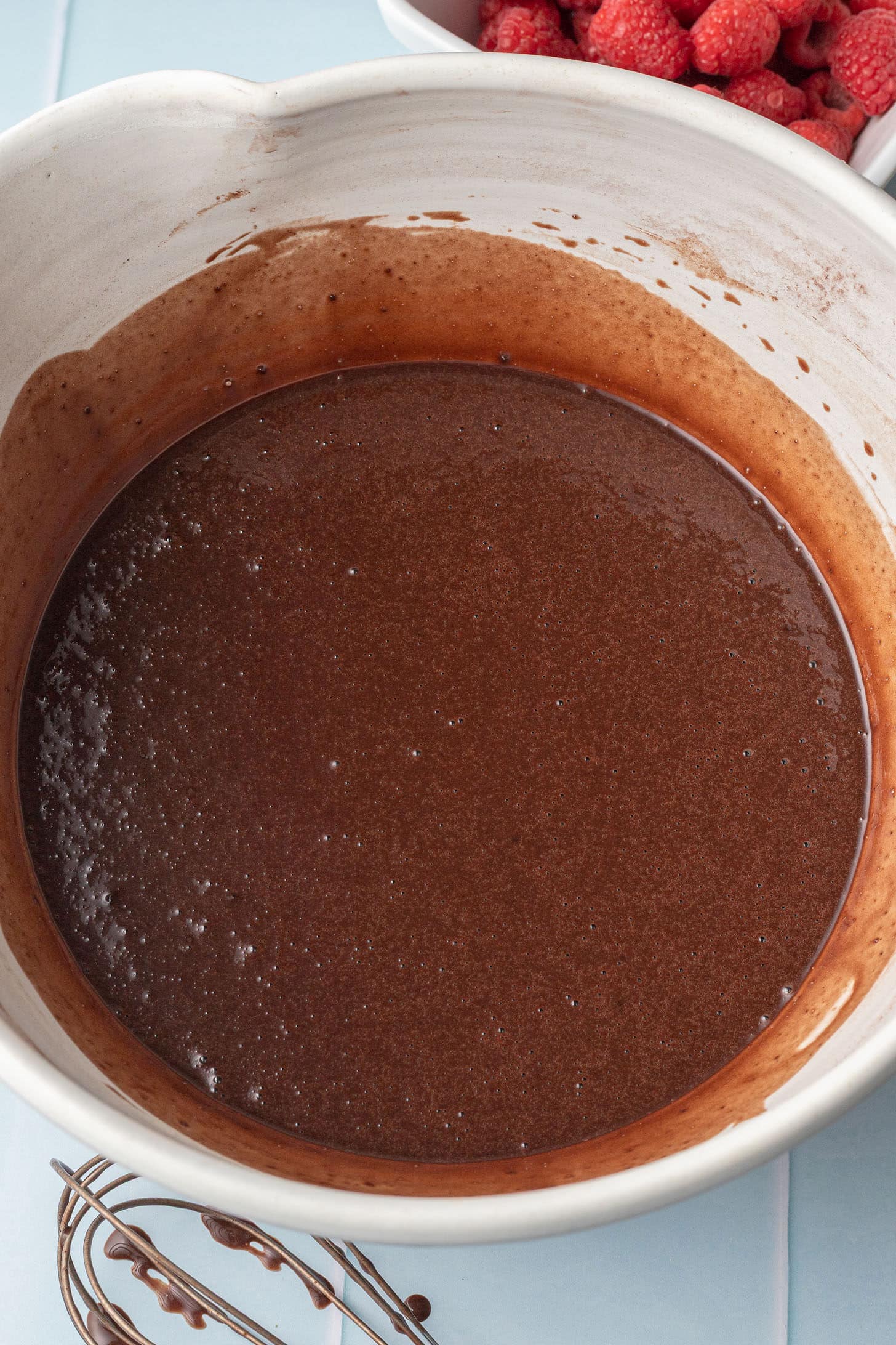 A white mixing bowl filled with smooth, glossy chocolate batter. A whisk with chocolate residue lies nearby. In the background, a bowl of fresh raspberries.