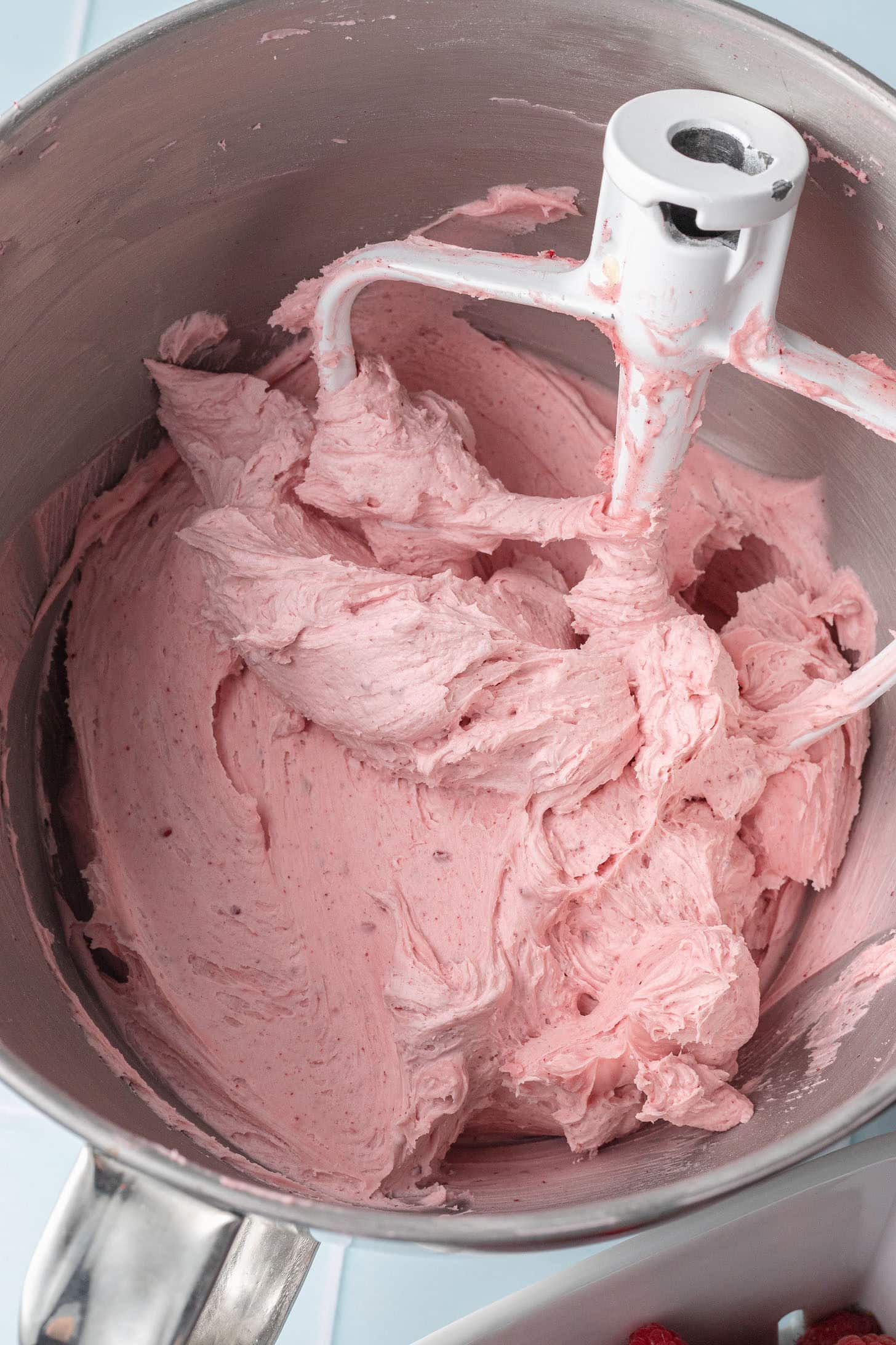 A stainless steel mixing bowl filled with fluffy, light pink frosting, showing creamy texture. Mixer paddle attachment partially visible.