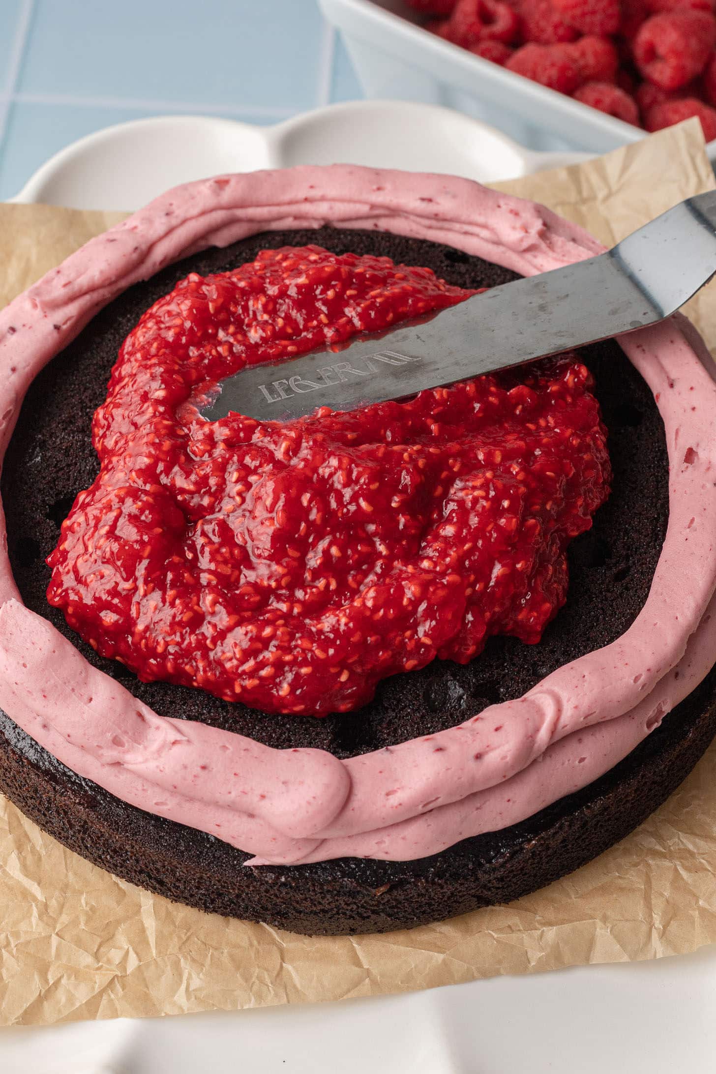 A chocolate cake with a pink frosting border and bright red raspberry topping being spread with a spatula. Fresh raspberries are visible in the background.