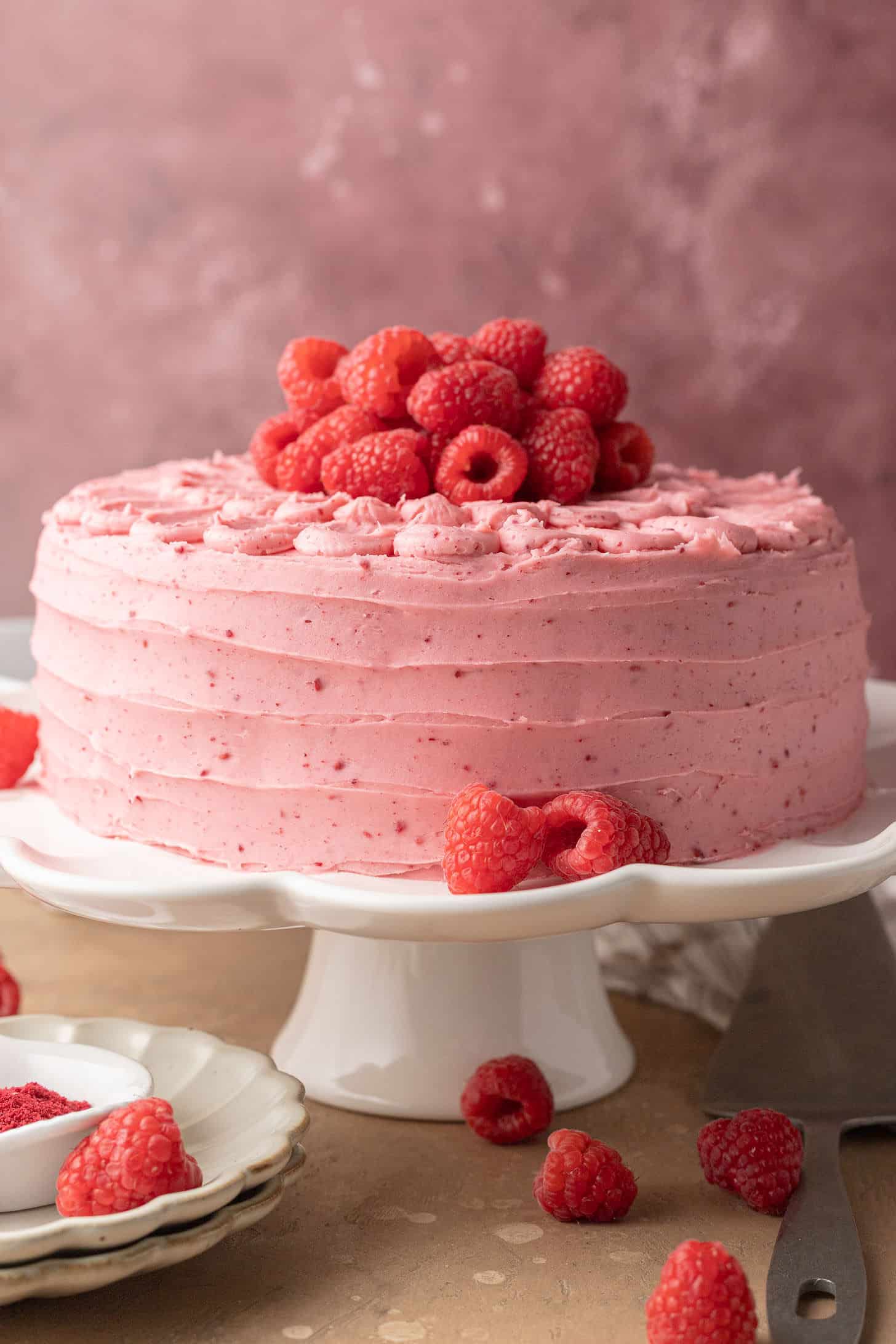 A pink frosted cake topped with fresh raspberries sits on a white pedestal against a pink textured background. Several raspberries are scattered around, conveying a fresh and inviting feel.
