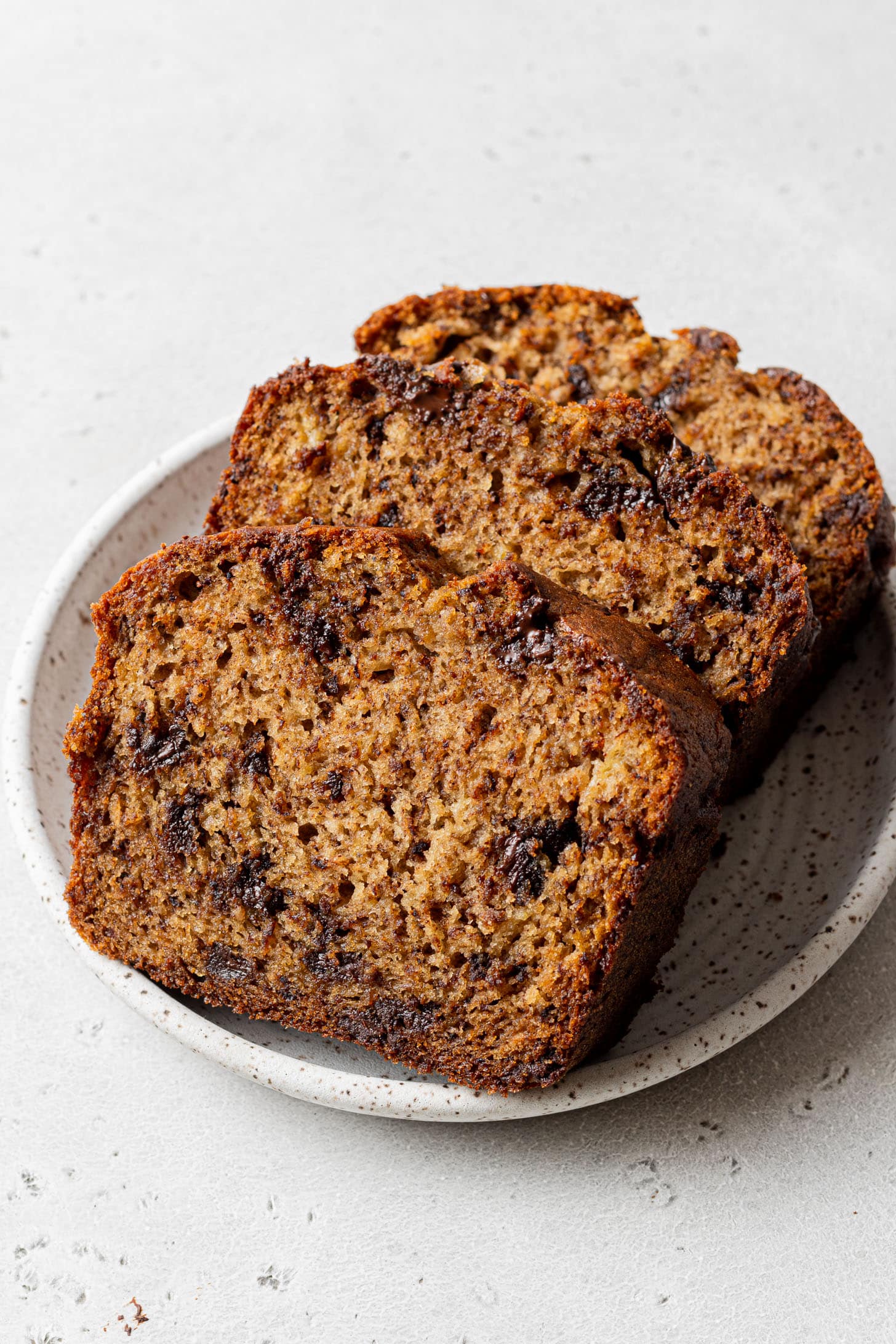 Three slices of moist banana bread with chocolate chips on a white, speckled plate. The bread appears soft and rich, creating a warm, inviting feel.