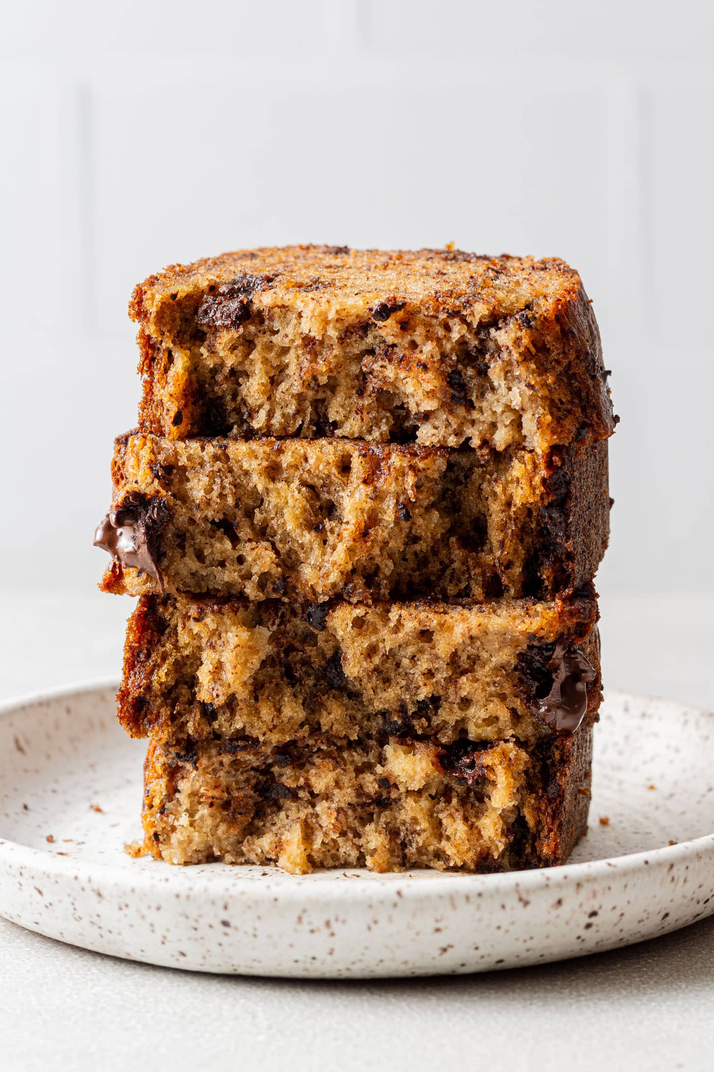Three slices of moist banana bread with melted chocolate chips stacked on a speckled white plate, evoking warmth and indulgence.
