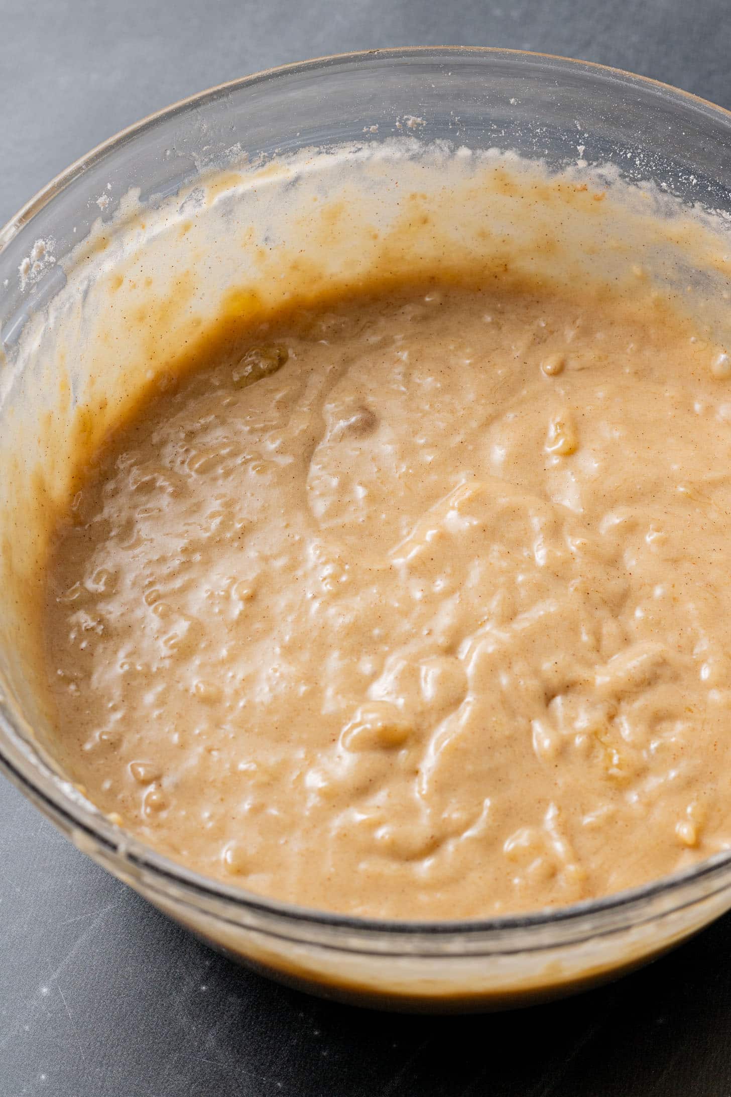A glass bowl filled with thick, creamy batter speckled with small banana chunks, set against a gray background, conveying a sense of anticipation for baking.
