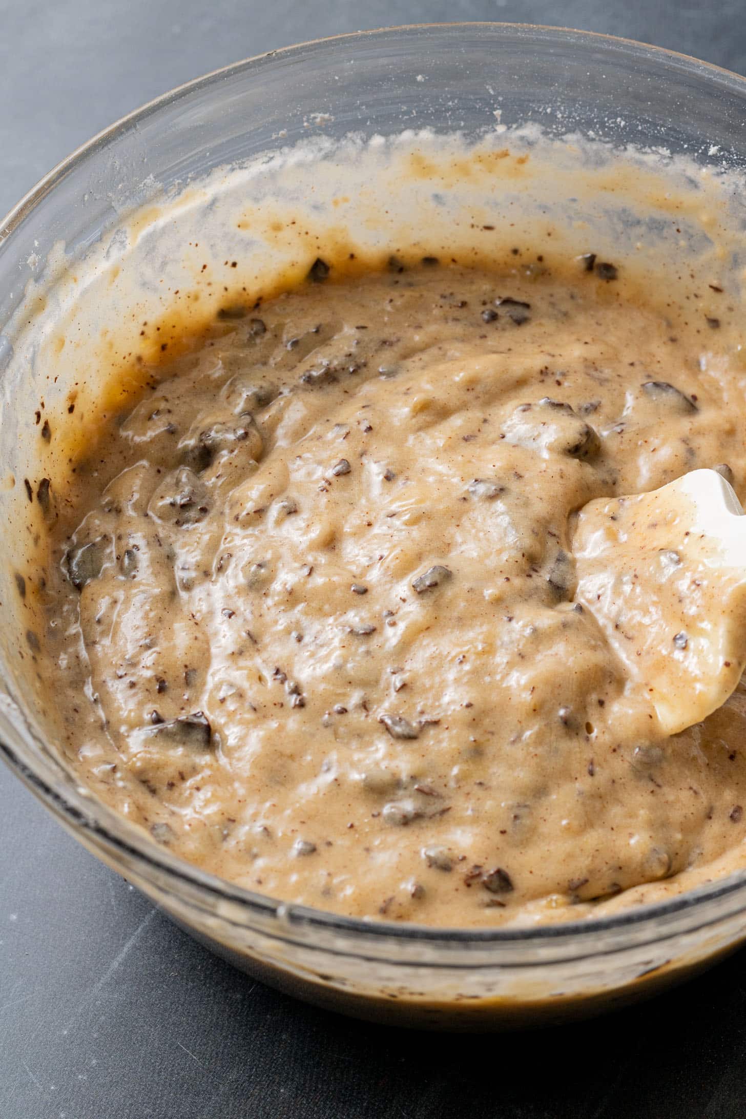 Close-up of a glass bowl filled with thick banana bread batter, speckled with chocolate chunks. A creamy texture is visible, suggesting a rich dessert.