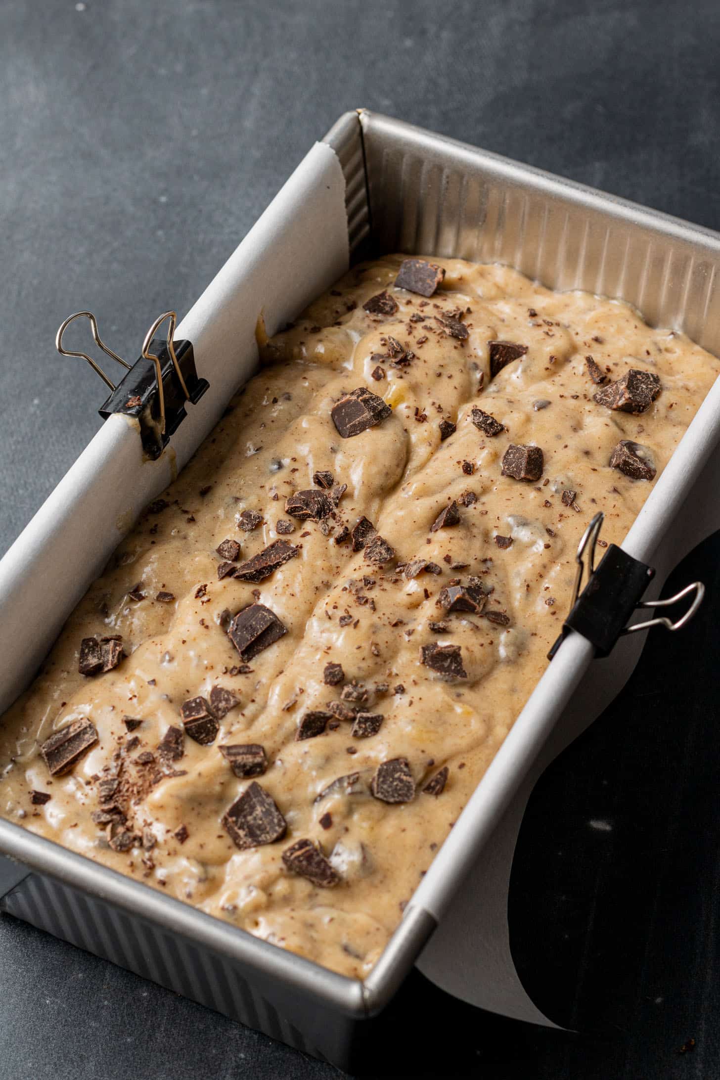Raw banana bread batter with chocolate chunks fills a rectangular loaf pan, lined with parchment paper and secured with binder clips.