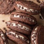 Chocolate sandwich cookies filled with cream and rolled in chocolate chips are stacked on brown parchment paper. A small bowl of chocolate chips is nearby.