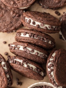 Chocolate sandwich cookies filled with cream and rolled in chocolate chips are stacked on brown parchment paper. A small bowl of chocolate chips is nearby.