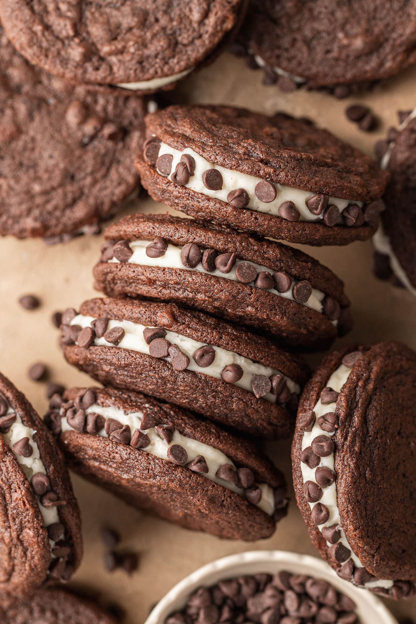 Chocolate sandwich cookies filled with cream and rolled in chocolate chips are stacked on brown parchment paper. A small bowl of chocolate chips is nearby.