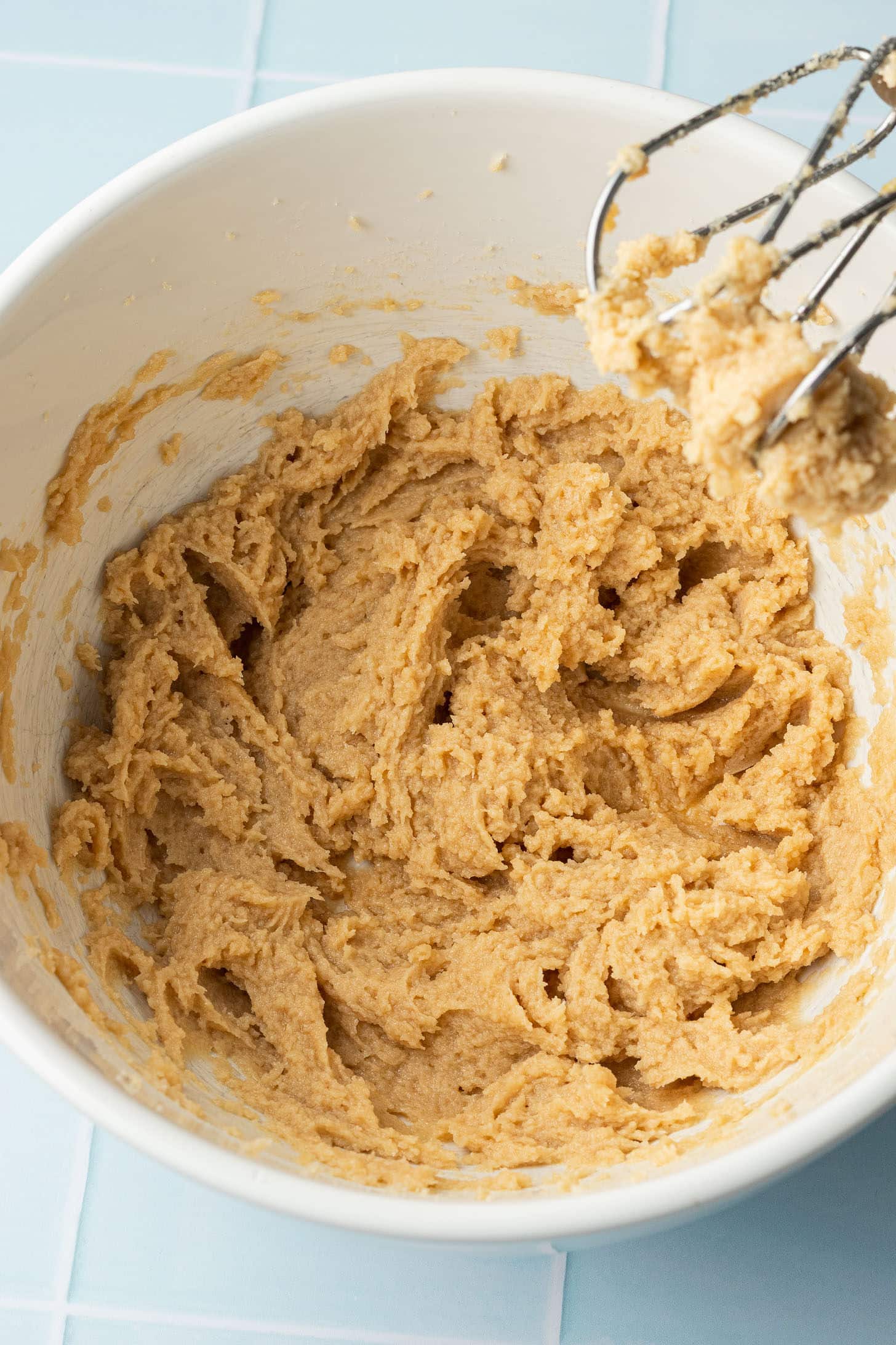 A mixing bowl containing a creamy, light brown cookie dough sits on a light blue tiled surface. A mixer with dough on its beaters hovers above the bowl.