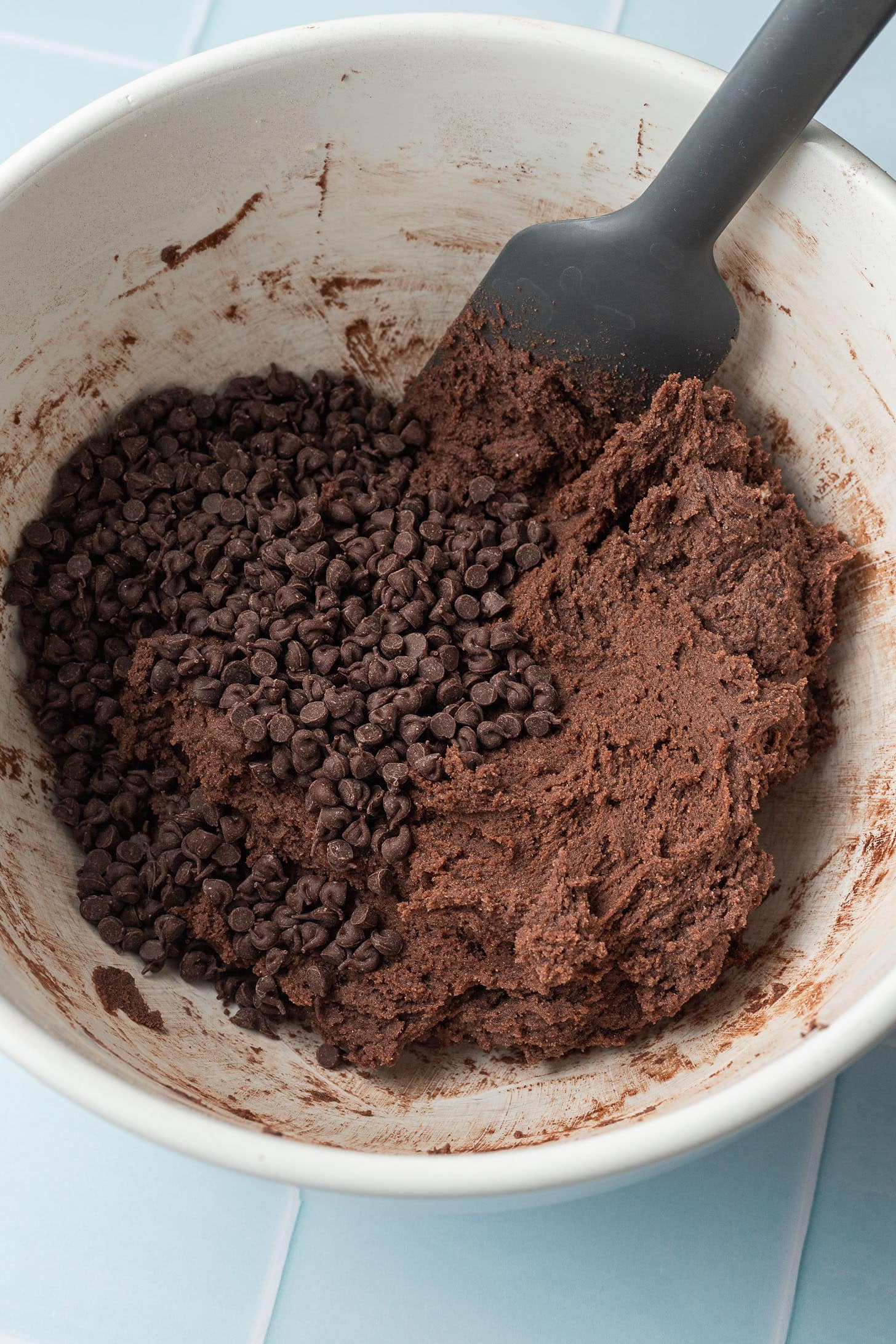 Chocolate cookie dough with mini chocolate chips in a white bowl. A black spatula is mixing the ingredients, creating a rich, indulgent tone.