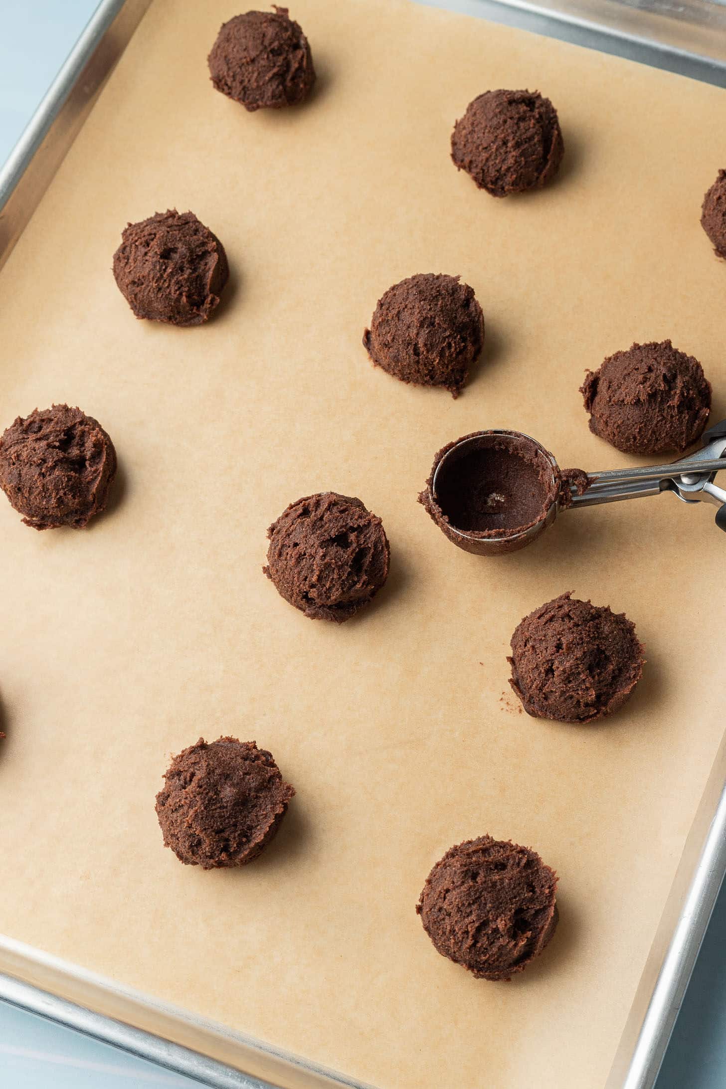 Scoops of chocolate cookie dough evenly spaced on a parchment-lined baking sheet, with a metal scoop resting nearby, ready for baking.