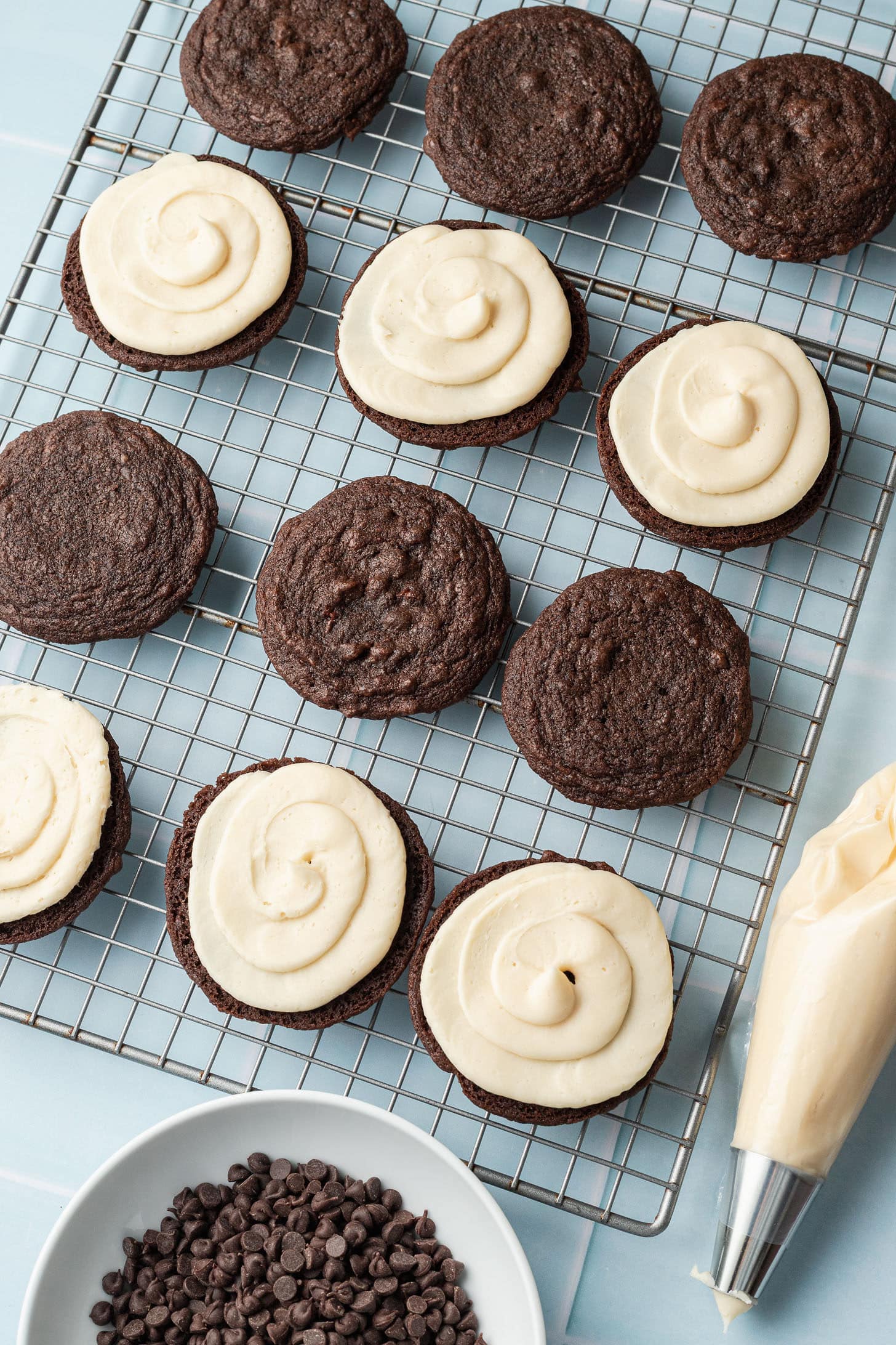 Chocolate cookies on a cooling rack, some topped with creamy white frosting. Nearby are a piping bag and a bowl of chocolate chips. Cozy and inviting.