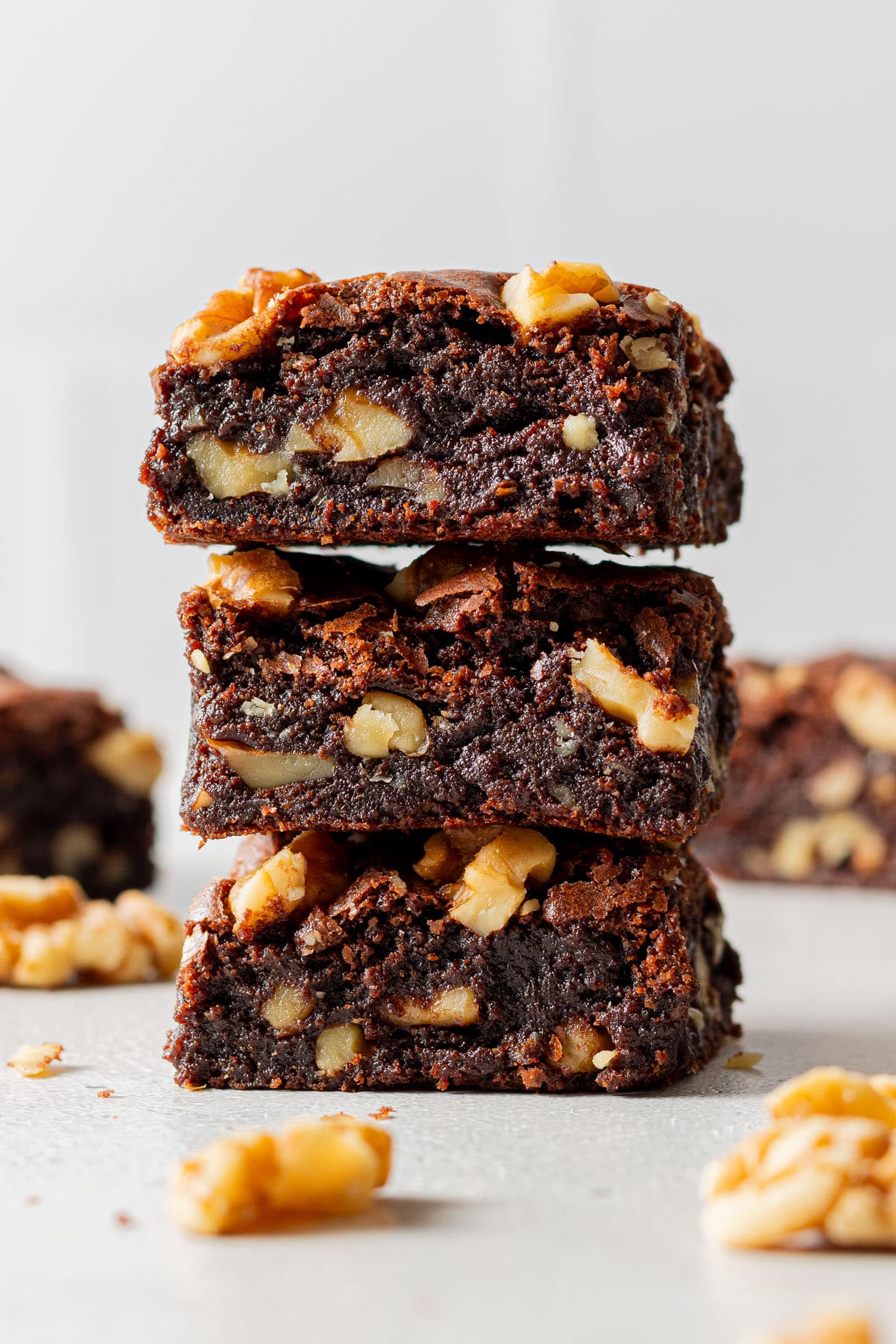 Stacked brownies with walnuts, richly chocolatey and moist, sitting on a light surface. Crumbs and walnut pieces scattered around add a homemade, inviting feel.