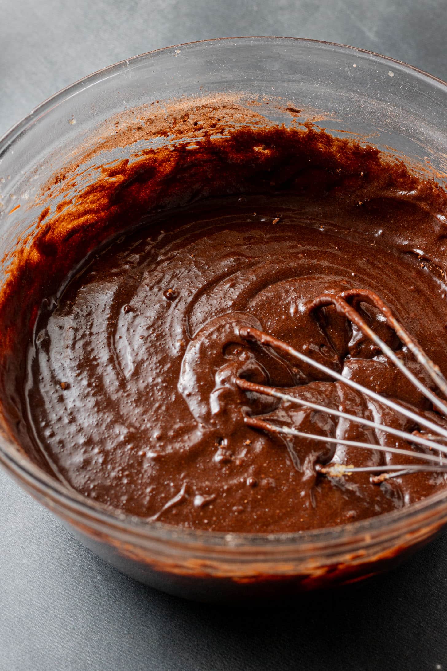 Chocolate cake batter in a glass bowl with a metal whisk, showing a smooth, rich texture. The scene conveys a sense of homemade baking.