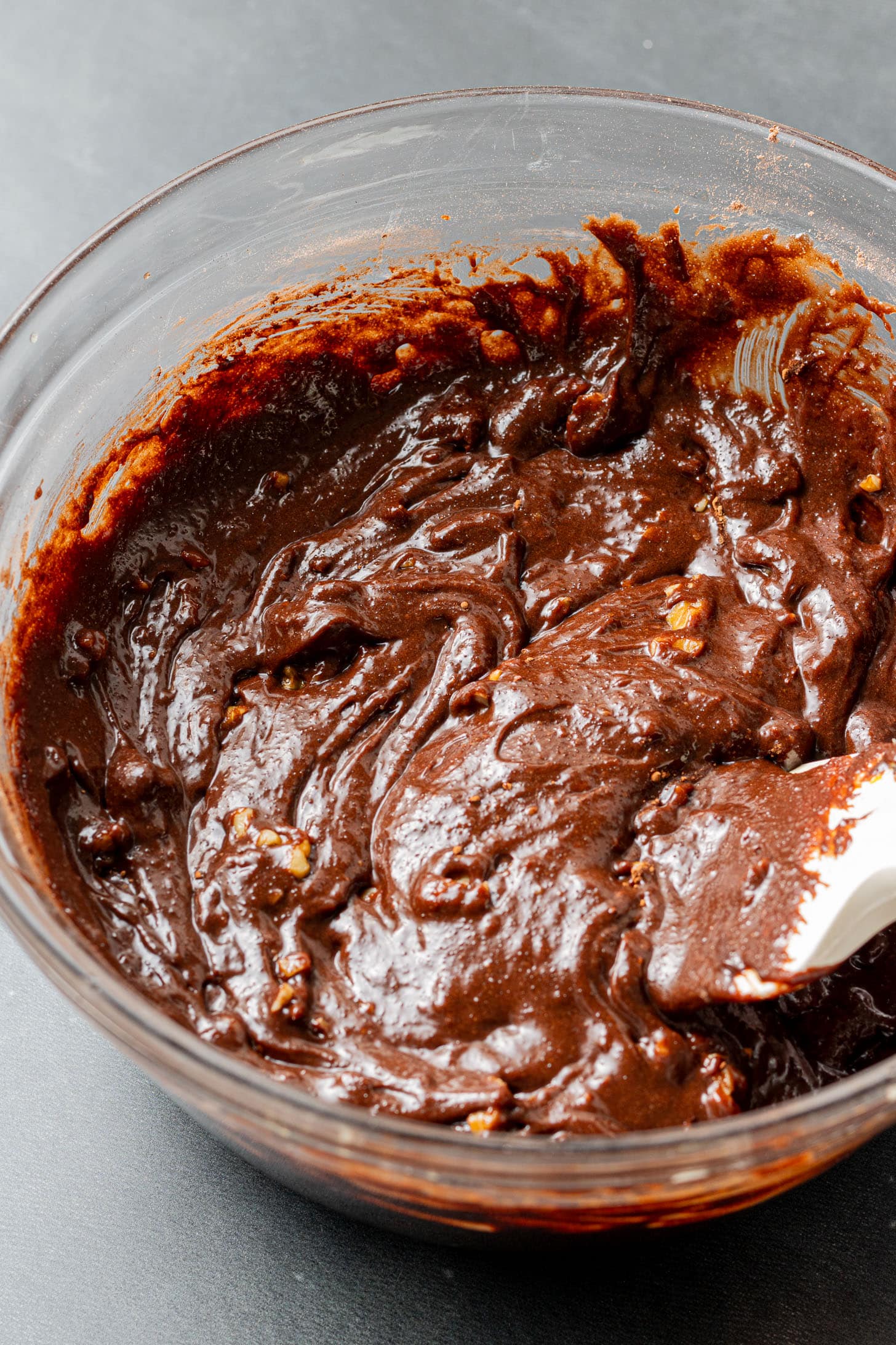 A glass bowl filled with rich, glossy brownie batter, mixed with visible chocolate chunks and nuts, sits on a dark surface, conveying a tempting, homemade feel.