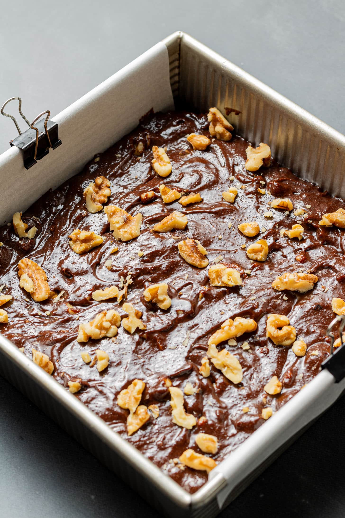 A square baking pan filled with rich, chocolate brownie batter topped with scattered walnuts rests on a gray surface, conveying a homemade, inviting feel.
