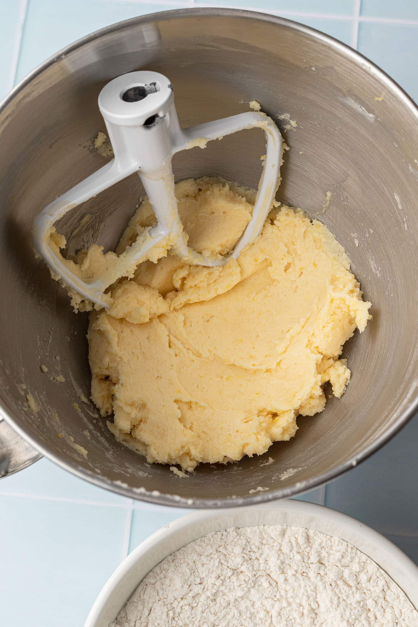 Mixing bowl with creamy yellow cookie dough and paddle attachment, on blue tiled counter. Nearby, a bowl of flour. Baking preparation scene.