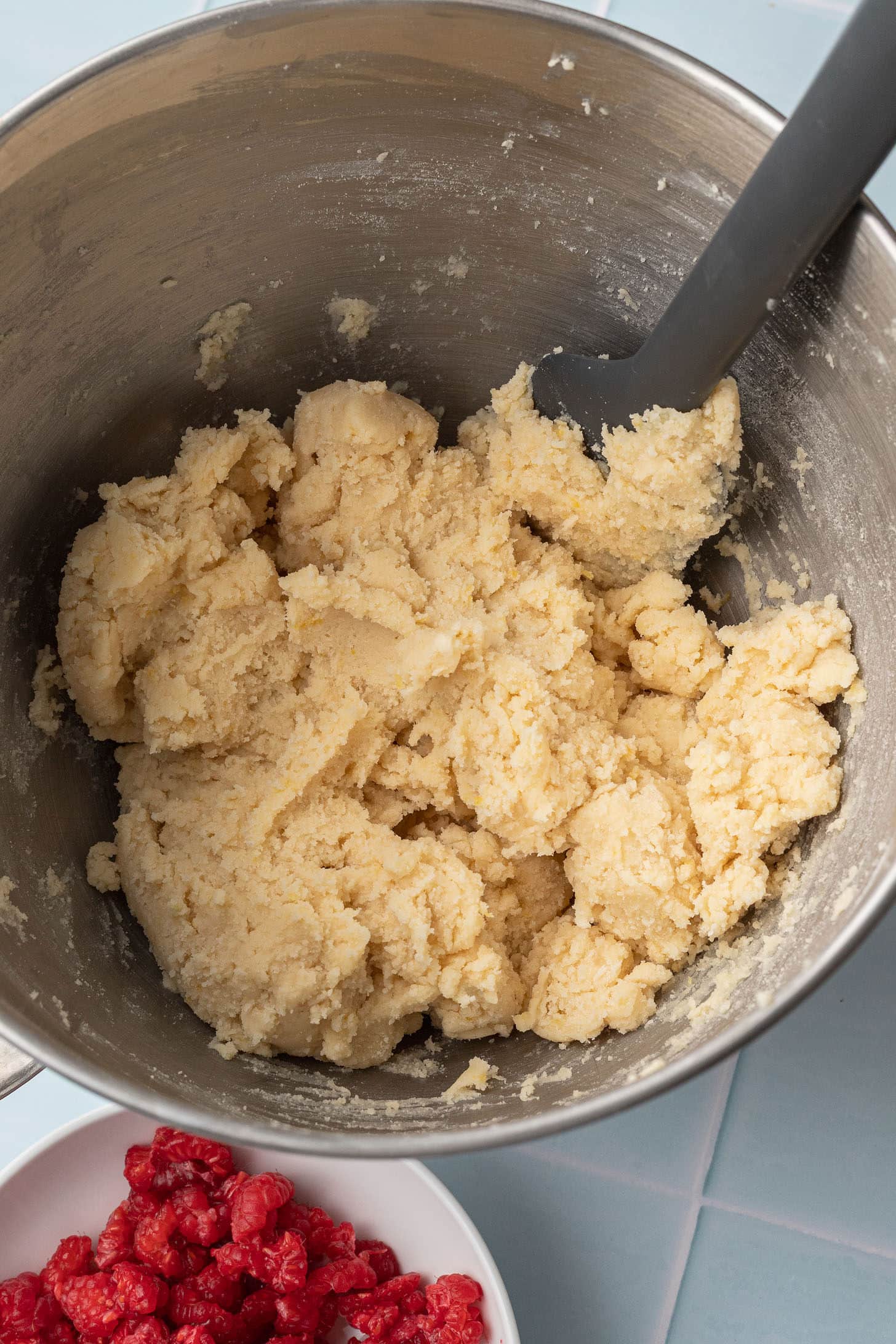 A stainless steel bowl contains crumbly, light beige dough. A gray spatula rests on the dough. Beside it, a bowl of fresh raspberries adds vibrant red color.