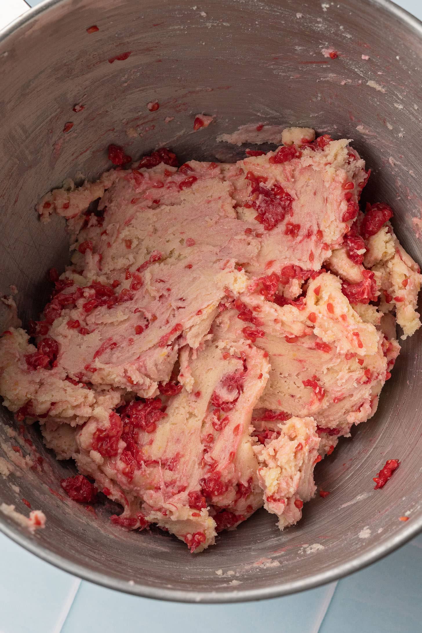 A metal mixing bowl filled with creamy cookie dough mixed with visible chunks of bright red raspberries, creating a textured and vibrant appearance.