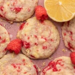 Close-up of raspberry lemon cookies on a pink surface. The cookies are studded with red raspberry pieces and lemon zest, with a bright lemon half and scattered raspberries, giving a fresh, vibrant feel.