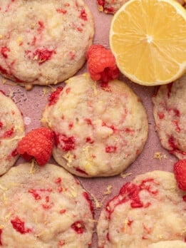 Close-up of raspberry lemon cookies on a pink surface. The cookies are studded with red raspberry pieces and lemon zest, with a bright lemon half and scattered raspberries, giving a fresh, vibrant feel.