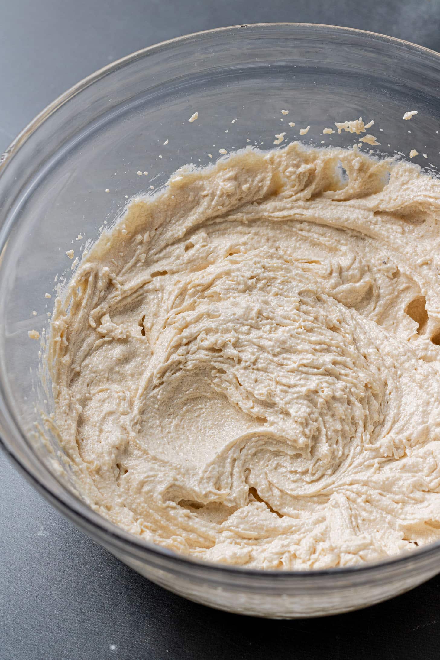 A glass bowl filled with smooth, creamy beige cookie dough batter sits on a dark countertop, suggesting a cozy and inviting baking scene.