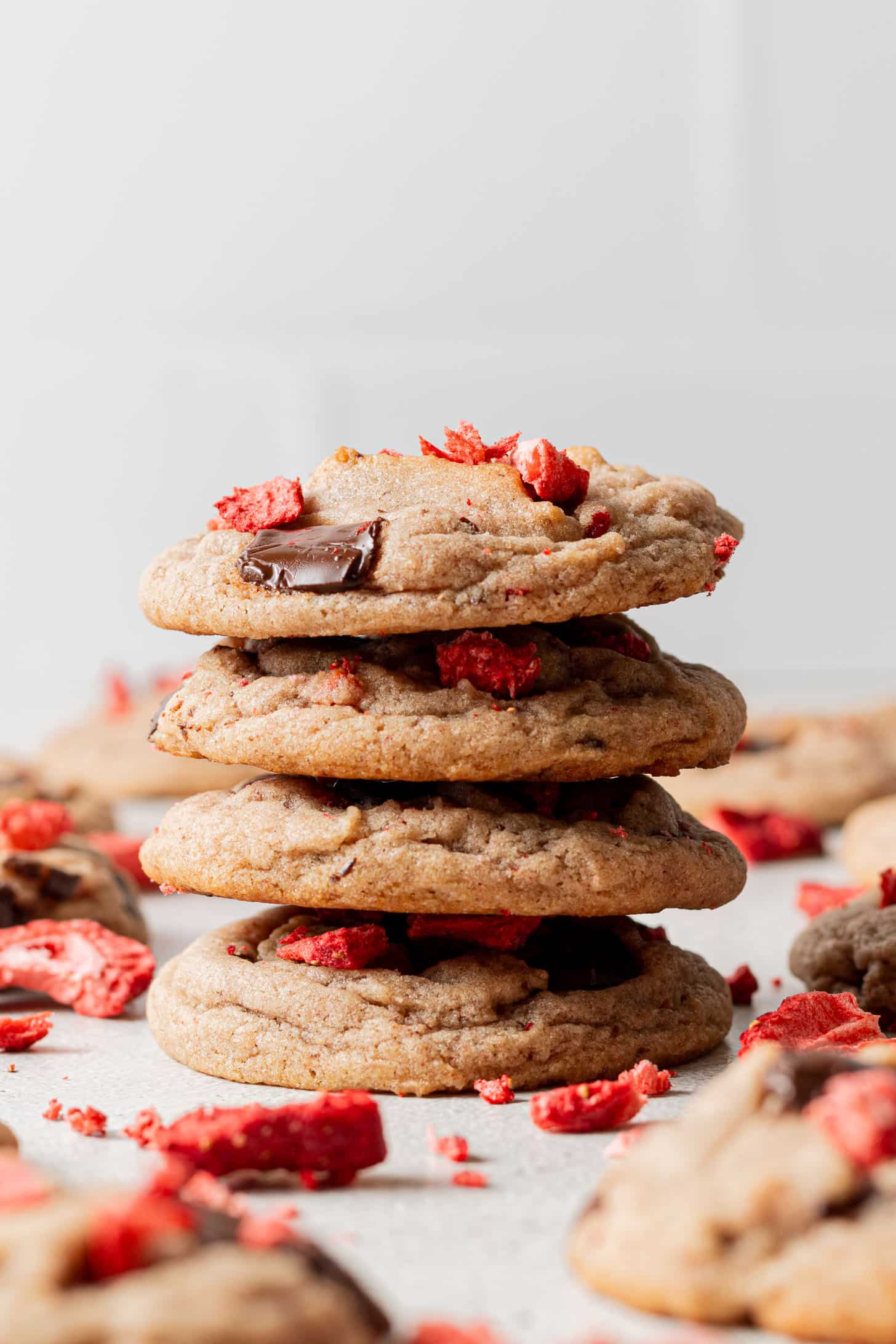 A stack of four cookies with chocolate chunks and red berry pieces on a light surface. Other cookies and scattered berry bits surround them, conveying a fresh, indulgent feel.