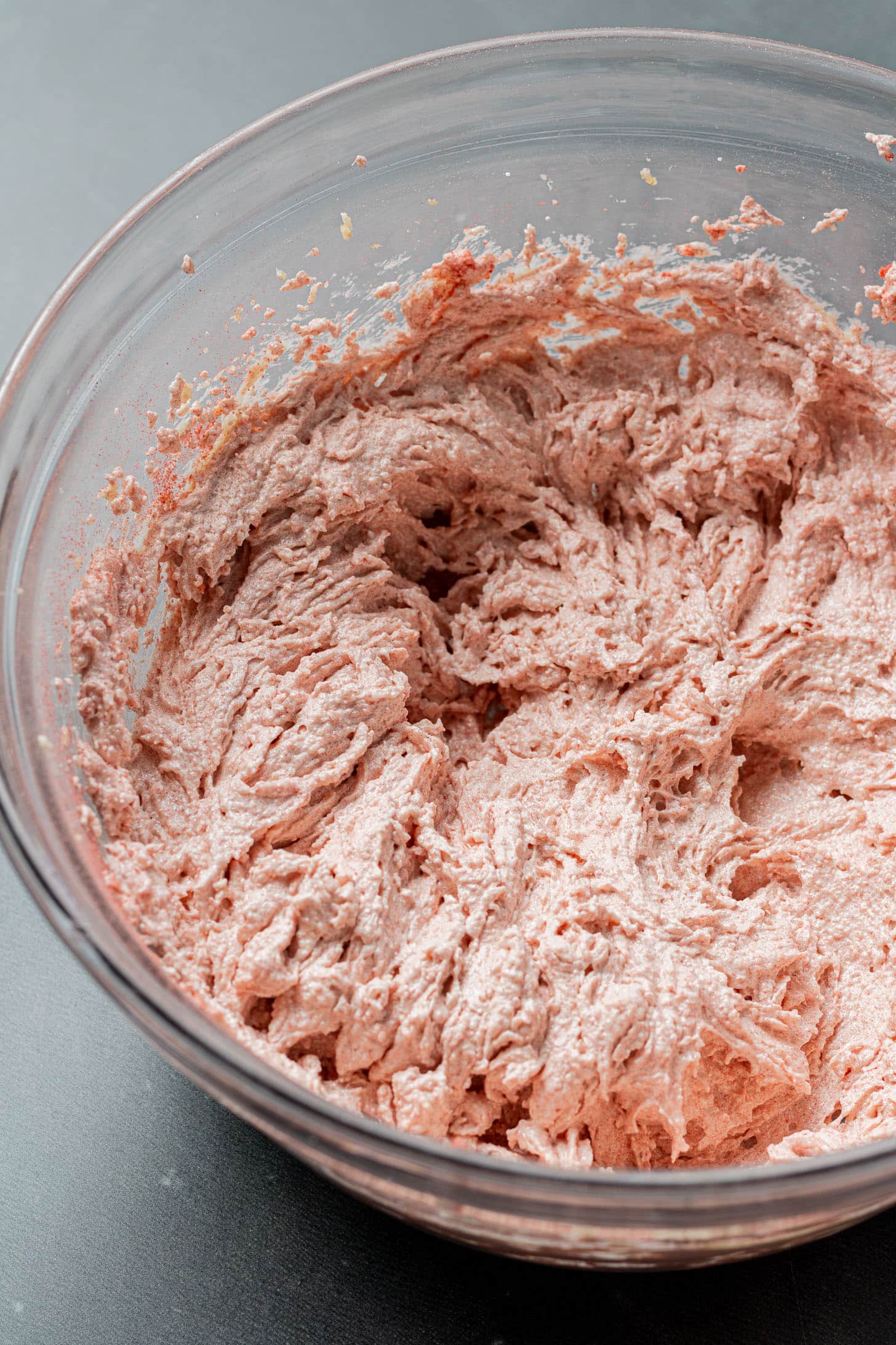 Creamy pink cake batter in a clear mixing bowl on a gray surface, showcasing a fluffy and smooth texture, indicating readiness for baking.