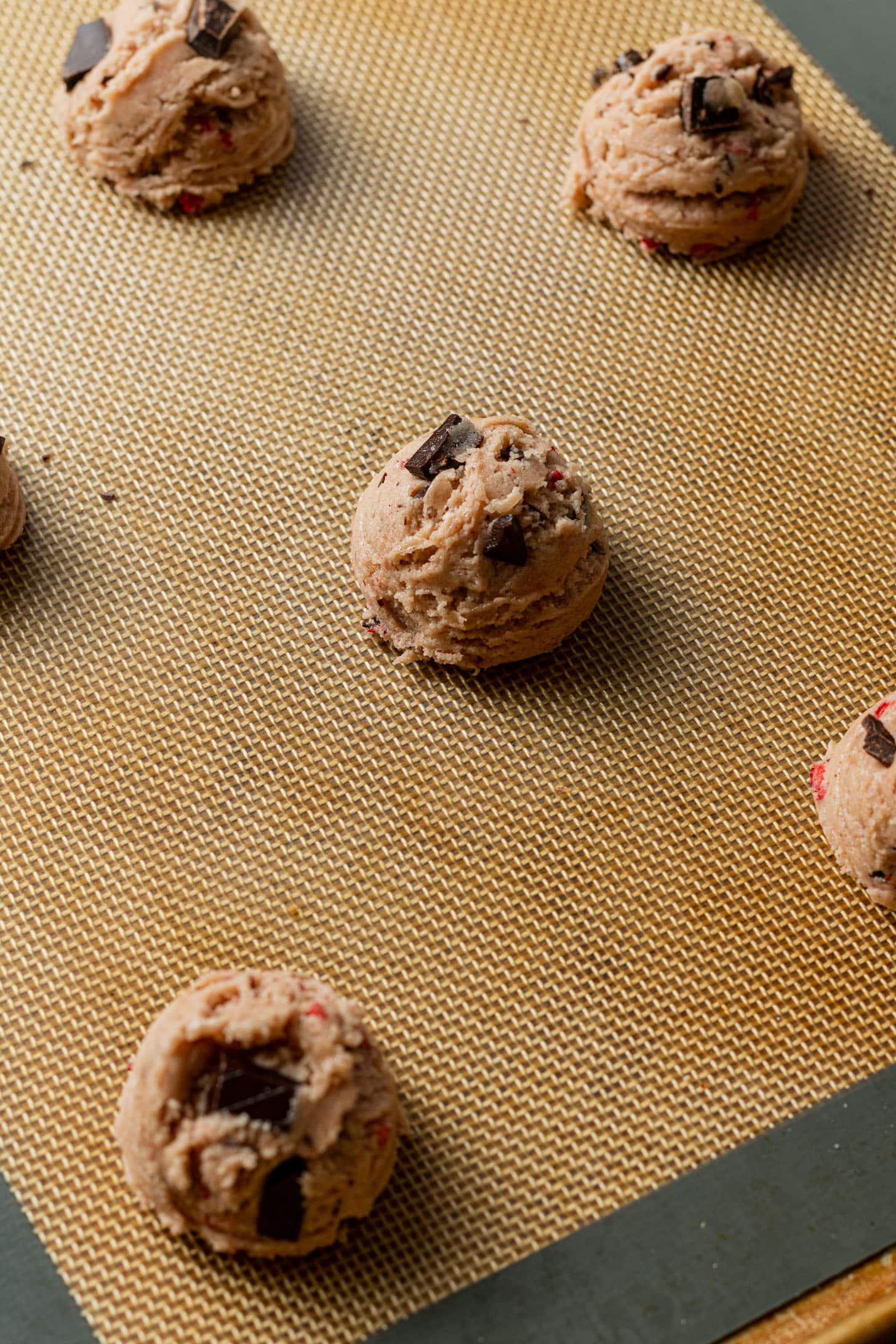 Scoops of chocolate chip cookie dough on a silicone baking mat, evenly spaced and ready for baking, convey a sense of anticipation and homemade coziness.