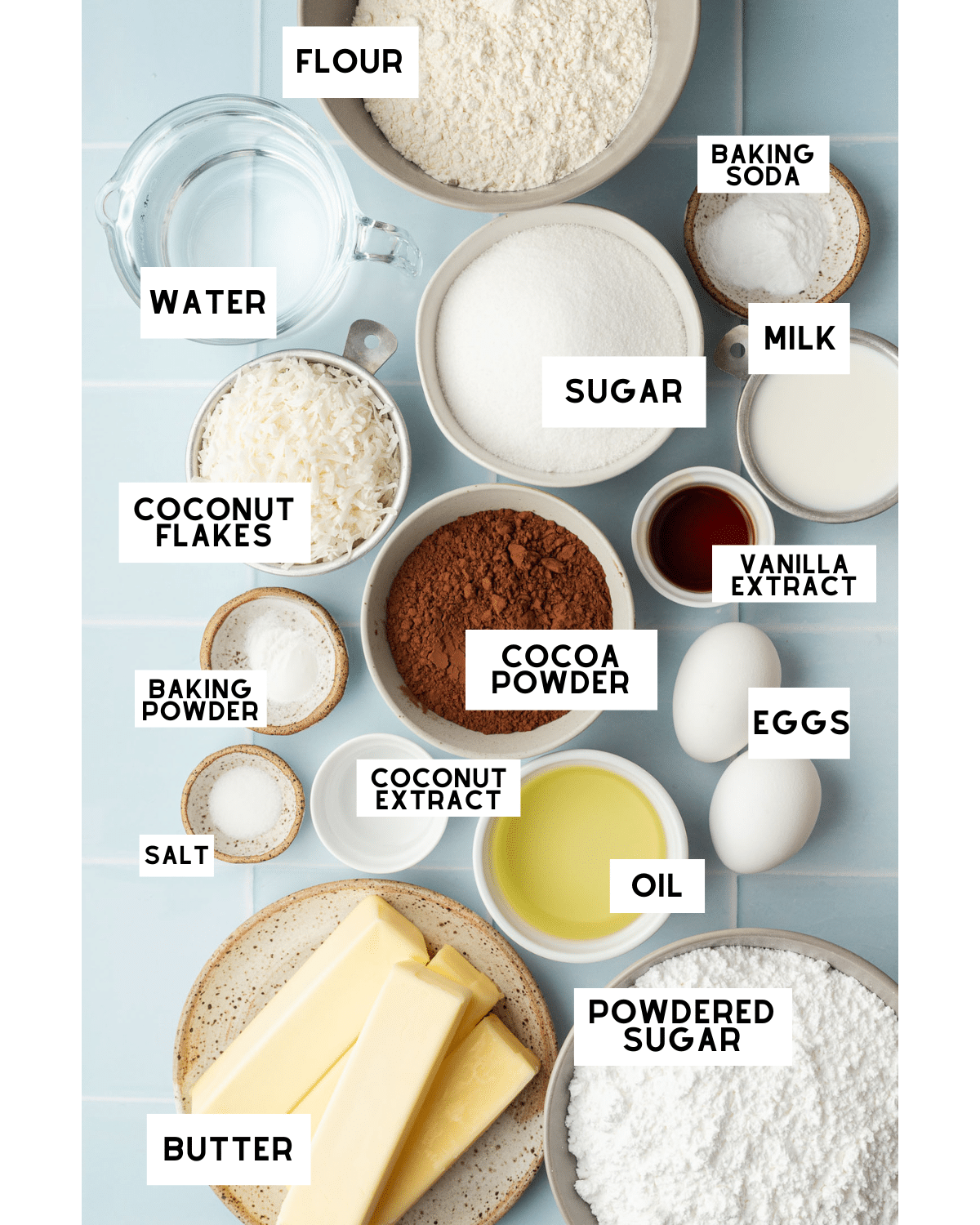 Flat lay of baking ingredients on a blue surface, including labeled bowls of flour, sugar, cocoa, coconut flakes, eggs, milk, and butter sticks.