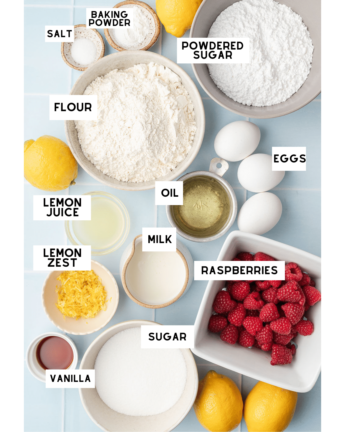 Ingredients for a raspberry lemon cake arranged on a blue surface: flour, eggs, raspberries, milk, lemon zest, lemon juice, oil, and sugar, with labels.