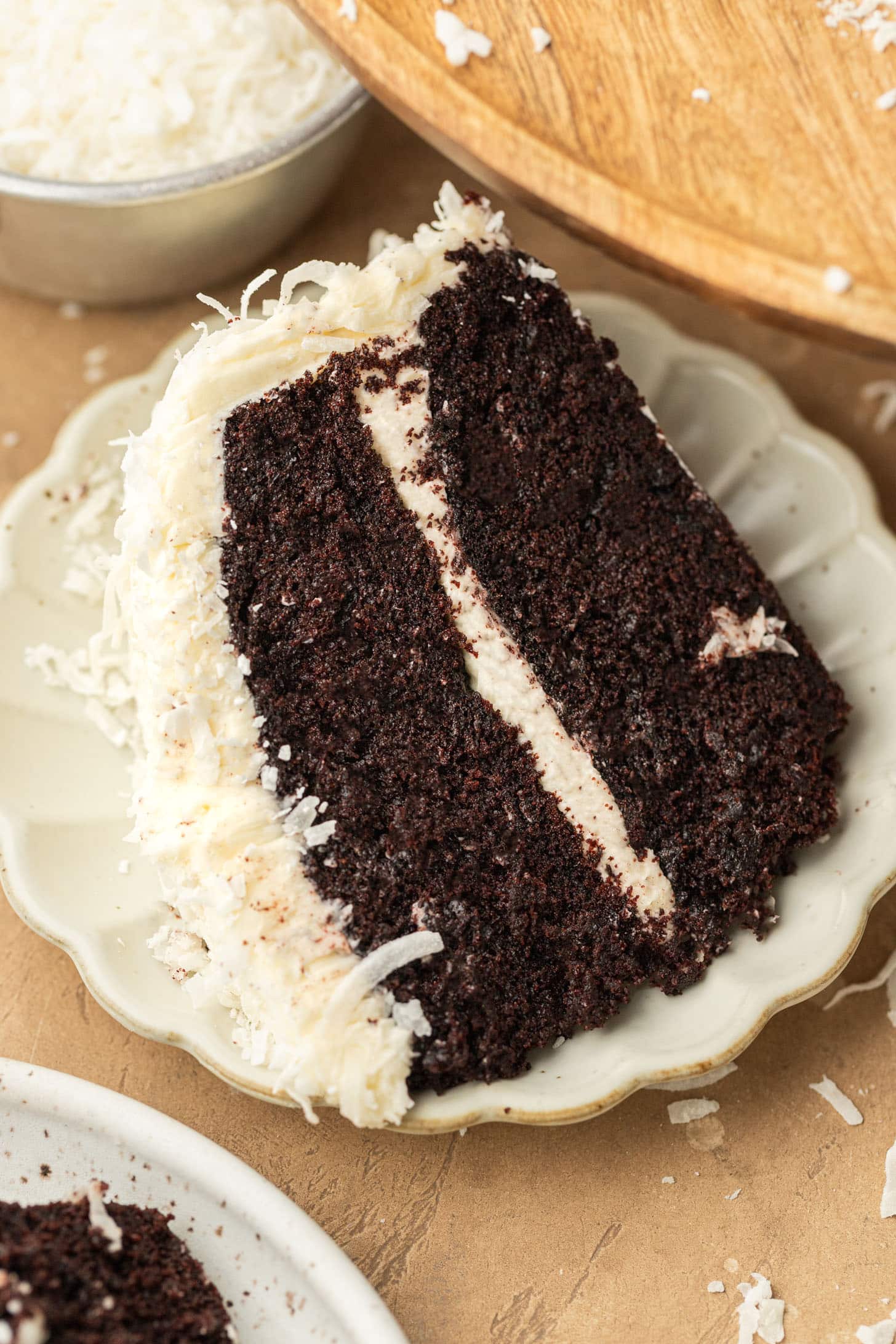 A slice of rich chocolate cake with creamy white frosting, topped with shredded coconut, sits on a decorative plate, conveying a tempting, indulgent mood.